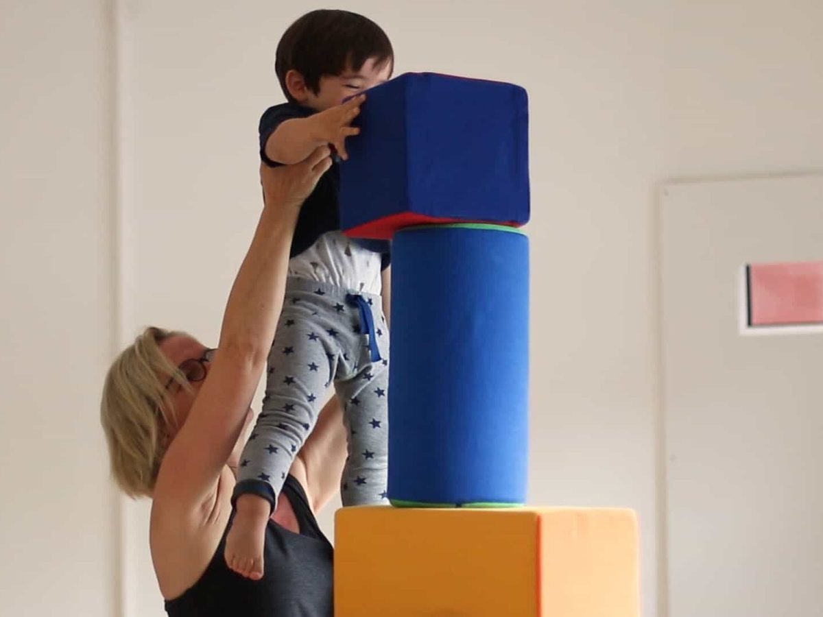 Eine Erwachsene hebt ein Baby, das nach einem blauen Schaumstoffwürfel auf einem Turm greift