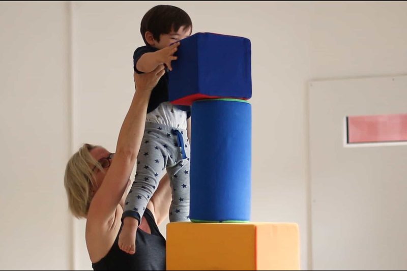 Eine Erwachsene hebt ein Baby, das nach einem blauen Schaumstoffwürfel auf einem Turm greift